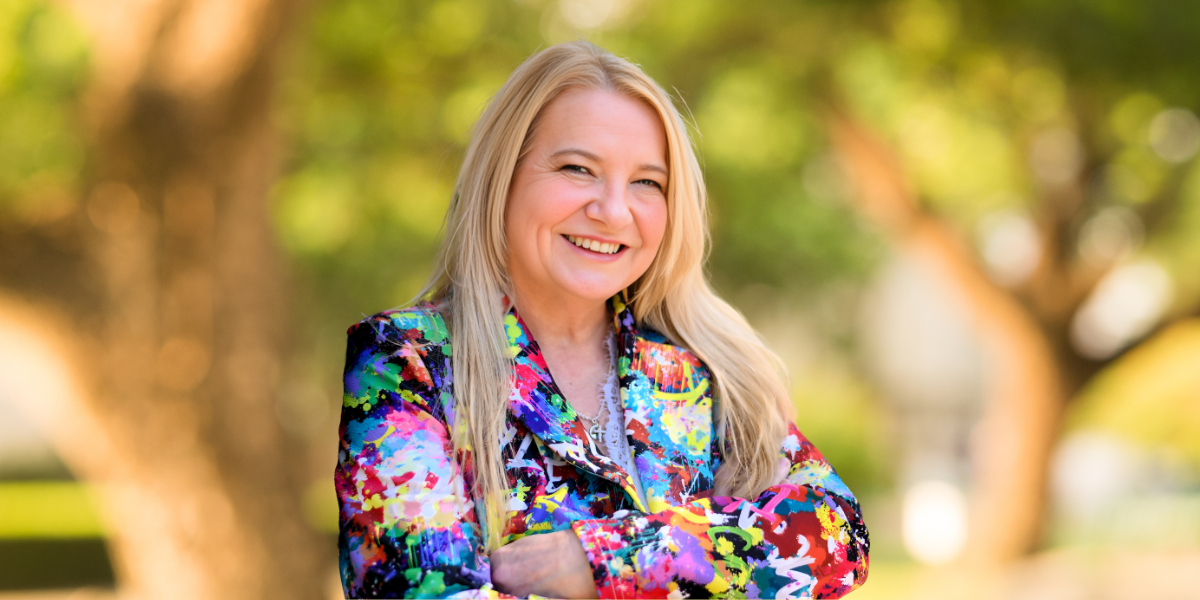 Smiling person with long blonde hair wearing a colorful abstract blazer outdoors
