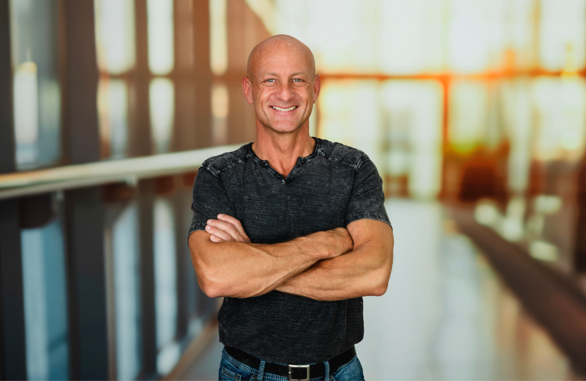 Smiling bald man with crossed arms posing in a blurred indoor setting