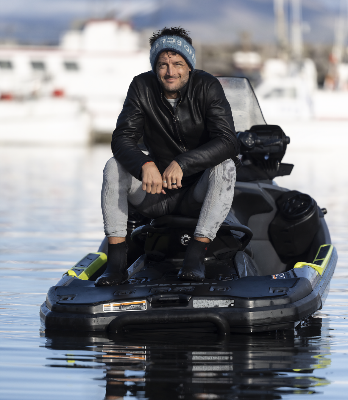 Man seated on a jet ski wearing a leather jacket and beanie on water