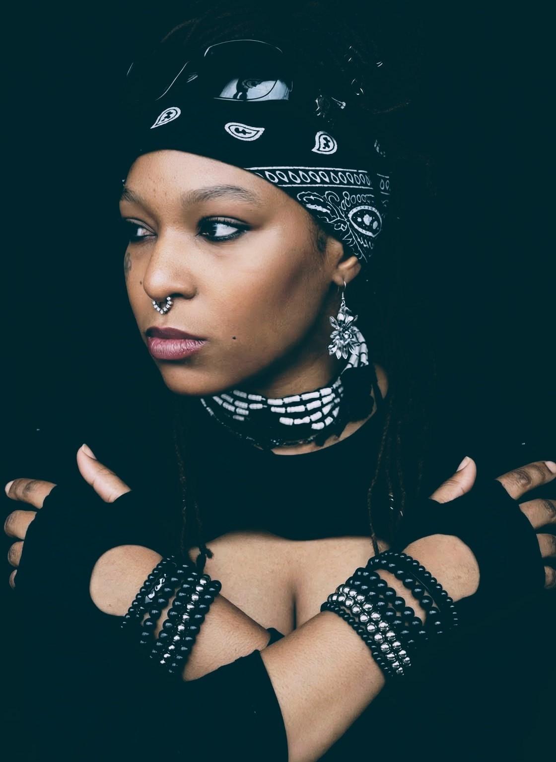 A woman with dreadlocks wearing black accessories and a bandana poses confidently.
