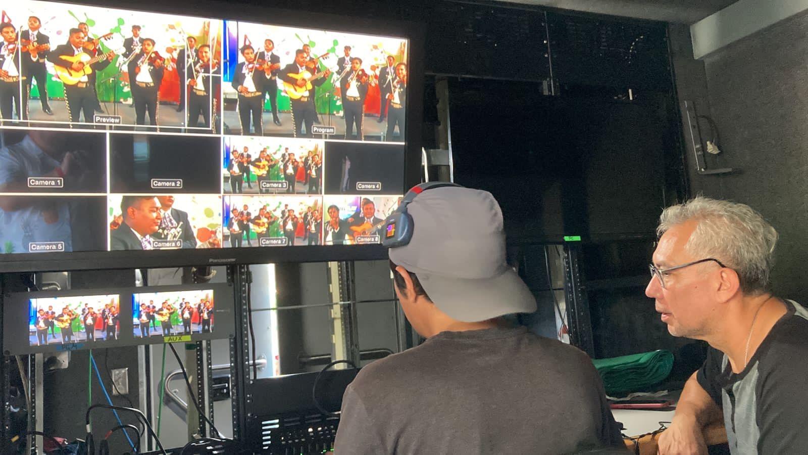 Two men observing multiple screens displaying live event footage inside a control room