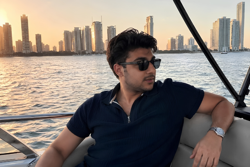 A young man in sunglasses relaxes on a boat with a city skyline at sunset