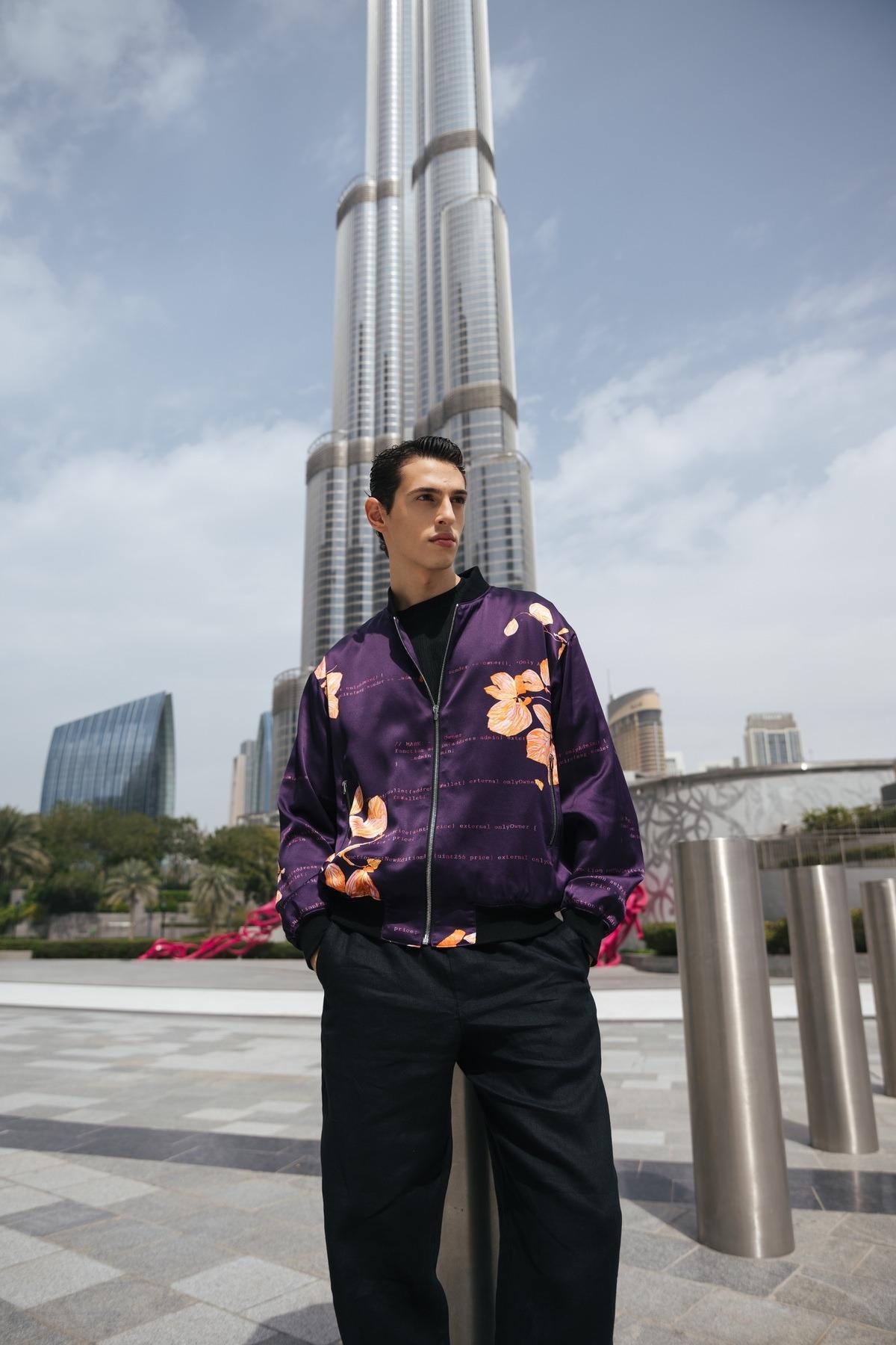 Man in floral purple jacket stands in front of a skyscraper under cloudy sky