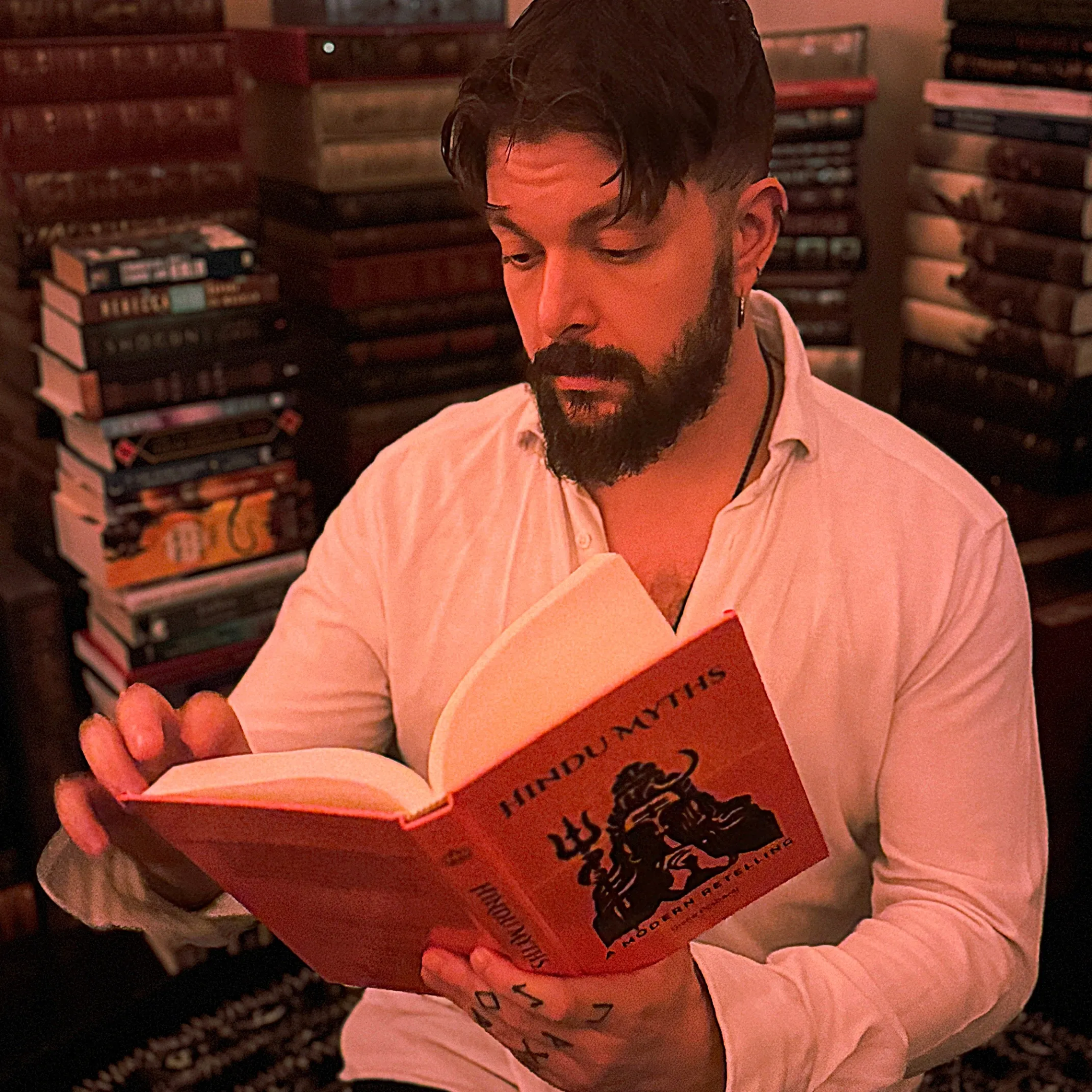 Man reading a book titled "Hindu Myths" among stacks of books in a cozy setting