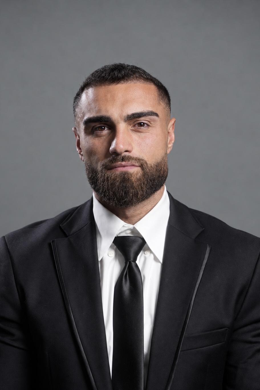 Man in a black suit and tie with a serious expression against a gray background