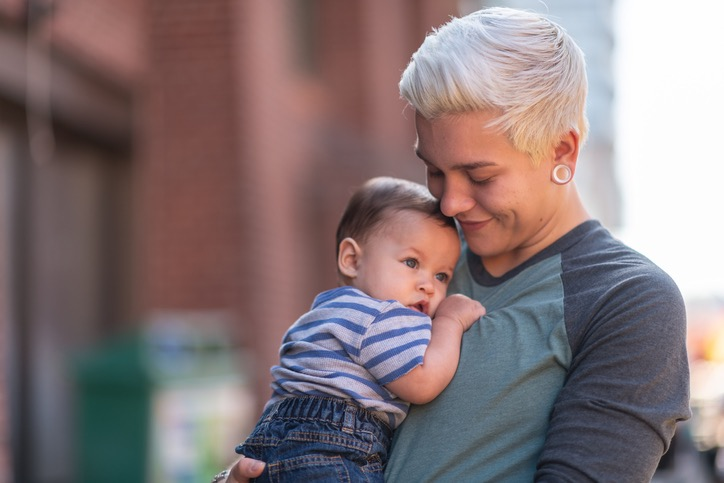 Person with short blonde hair holding a baby in a casual setting outdoors