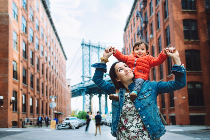 A woman lifts a child on her shoulders near a bridge and brick buildings