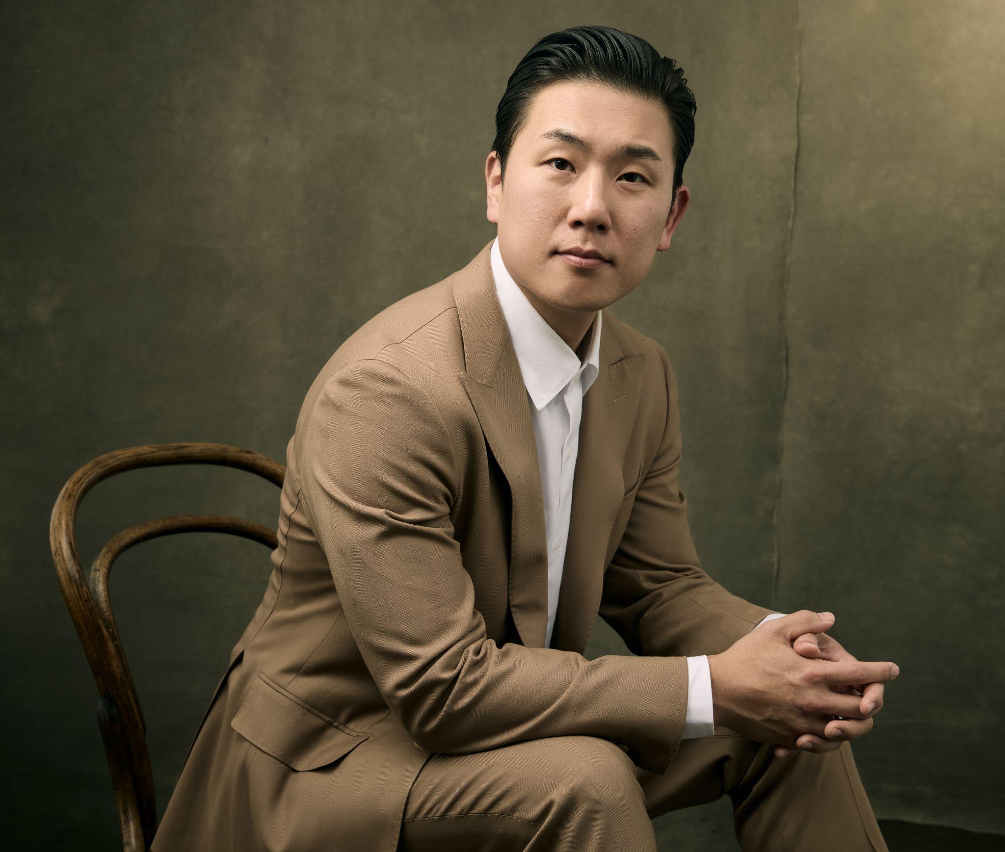A seated man in a tailored beige suit poses thoughtfully against a textured background