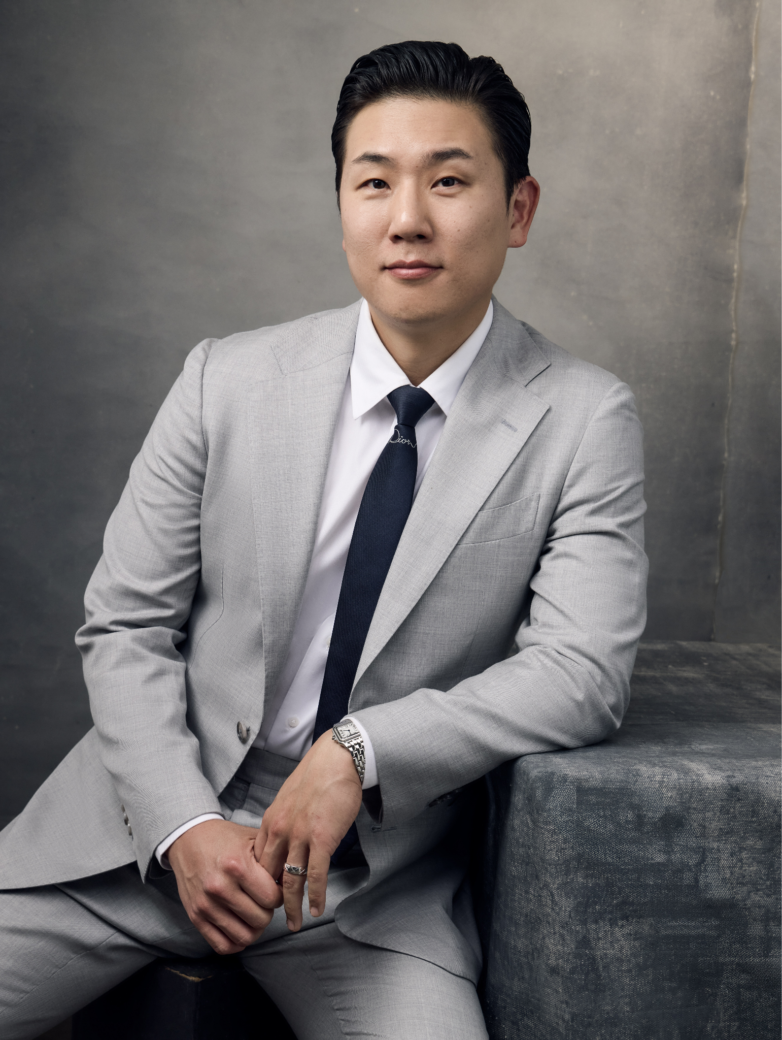 Man in a light gray suit poses confidently, sitting on a textured surface