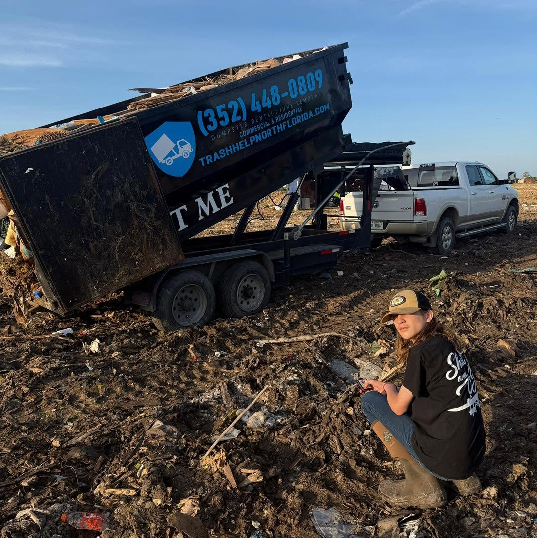 Person kneeling by a dumpster at a waste site surrounded by debris