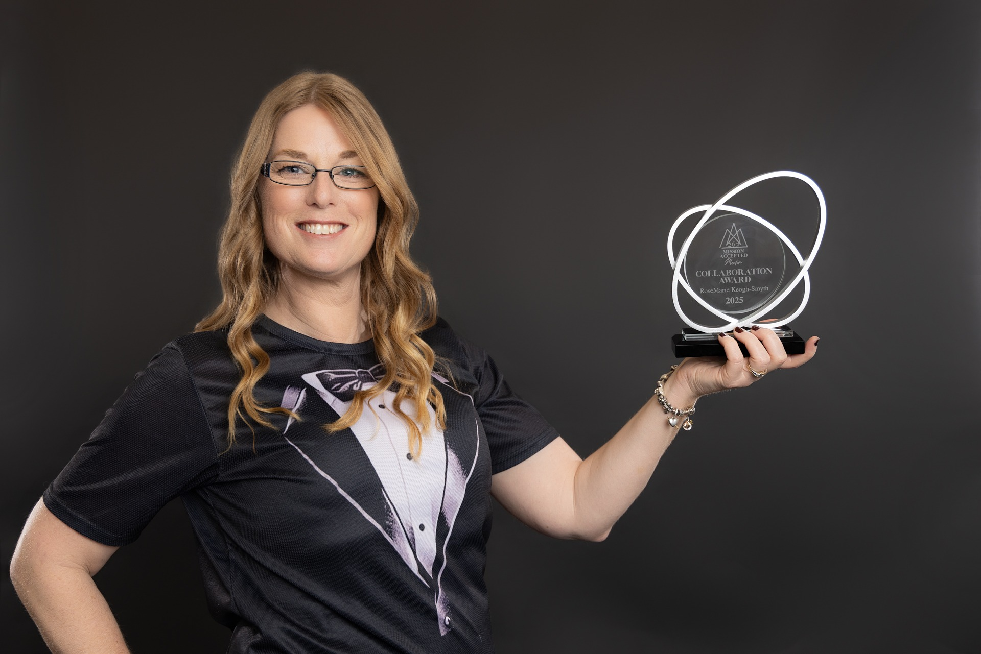 Smiling woman in a tuxedo t-shirt holding an award against a dark background