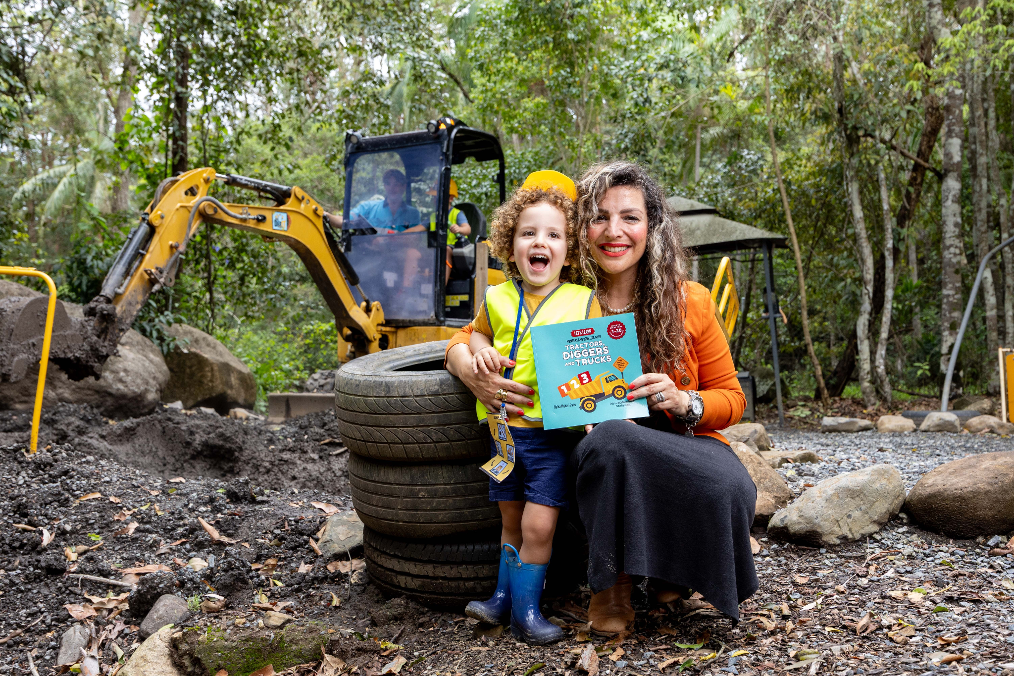 A woman and a child pose with a book near construction equipment in a forest.