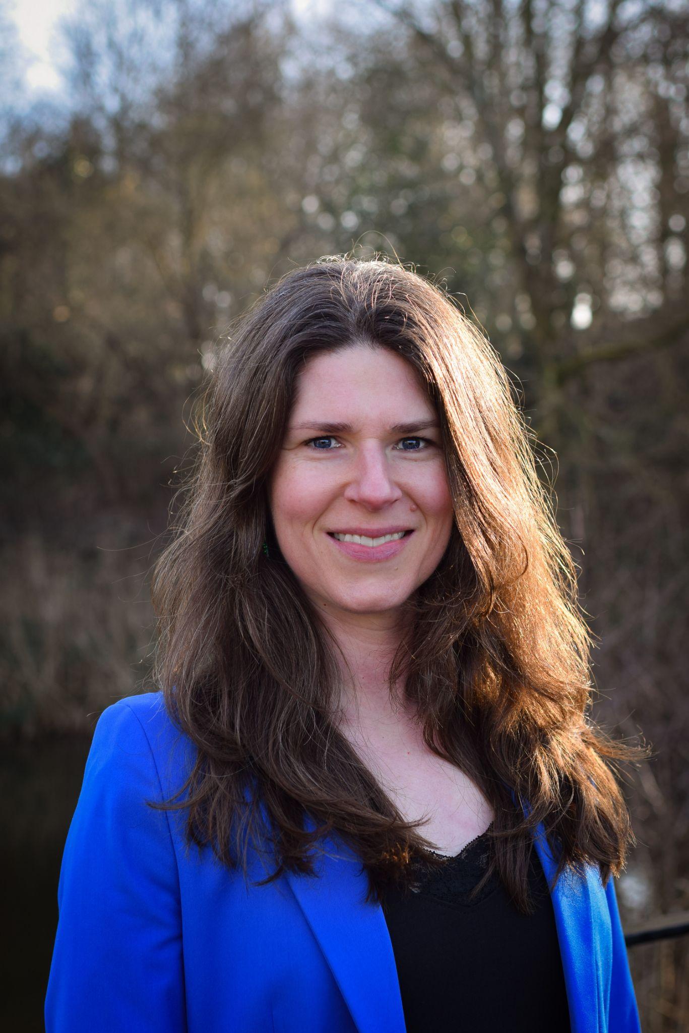 A woman with long hair smiles, wearing a blue blazer in a natural setting