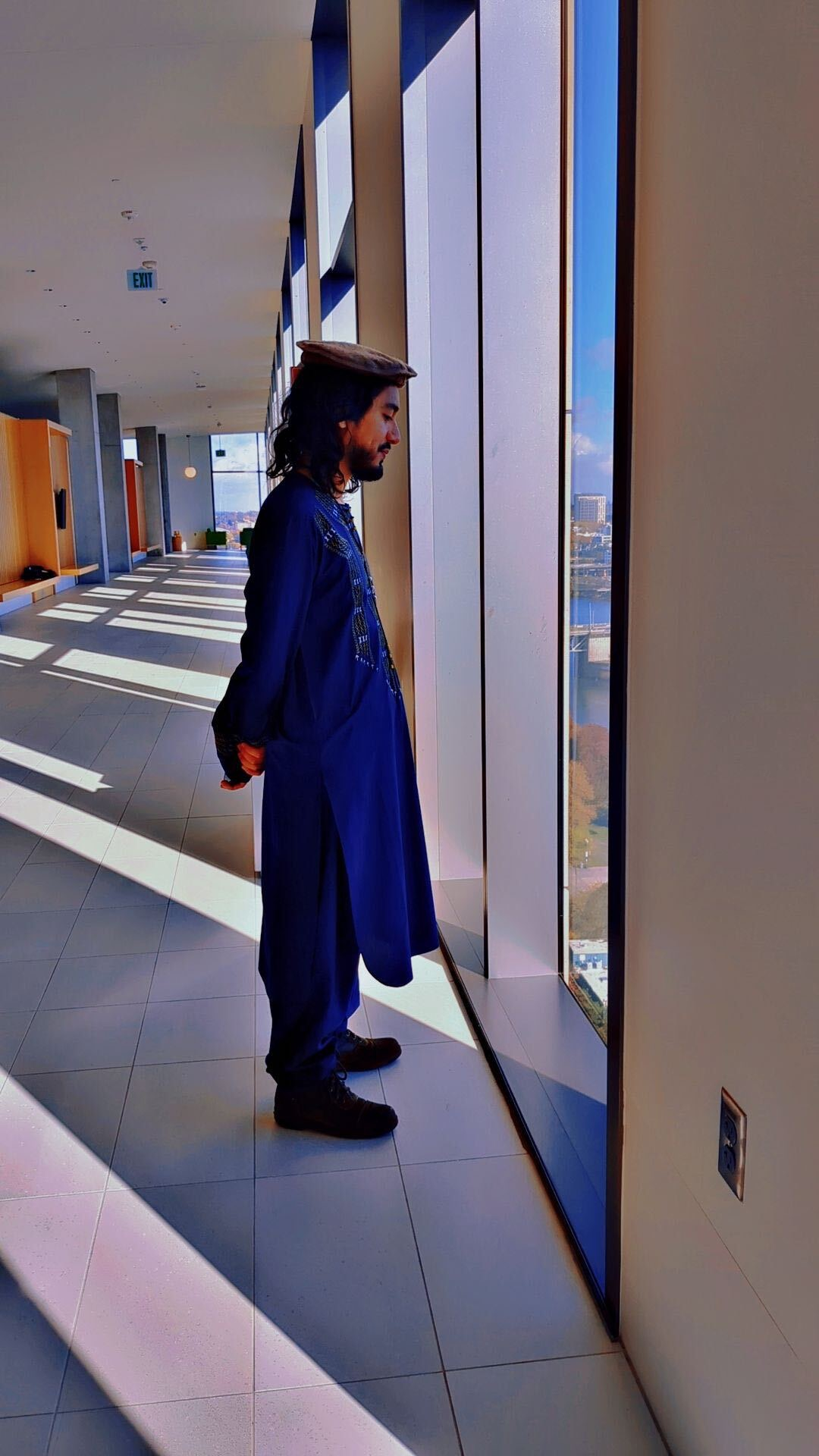 Man in traditional attire standing by a large window, gazing outside.