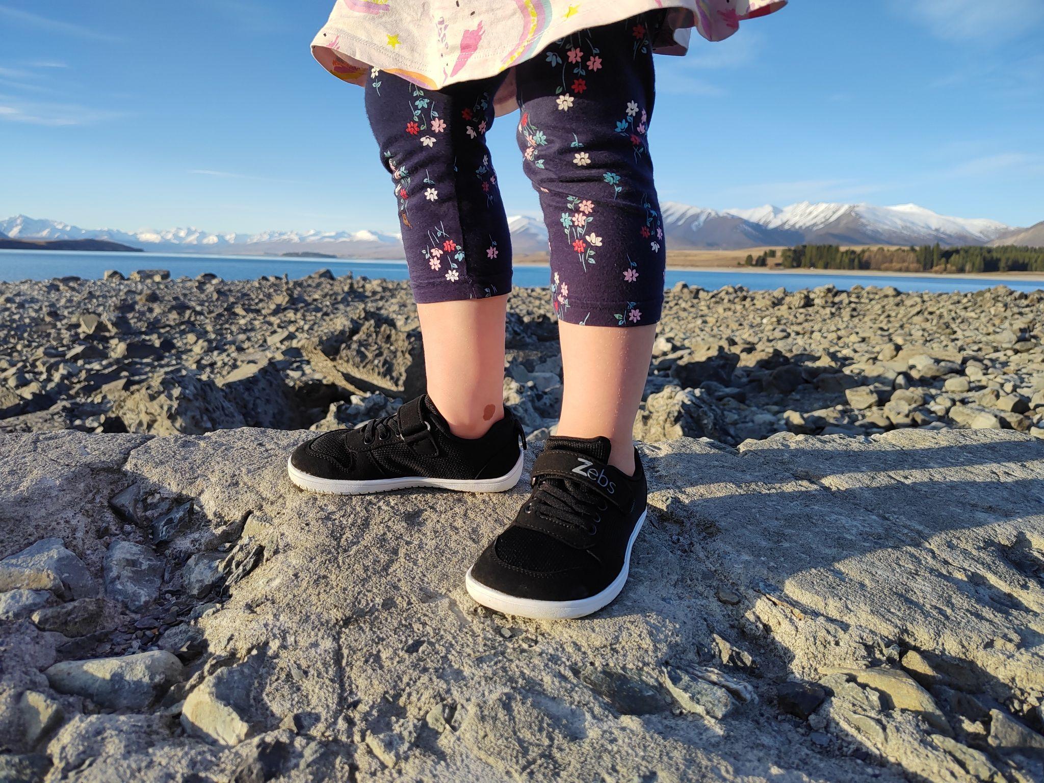 Child's legs in black sneakers standing on rocky shoreline with mountains in background