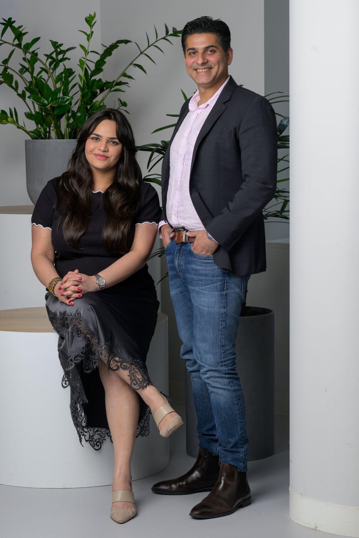A man and woman pose together in a modern indoor setting with plants.