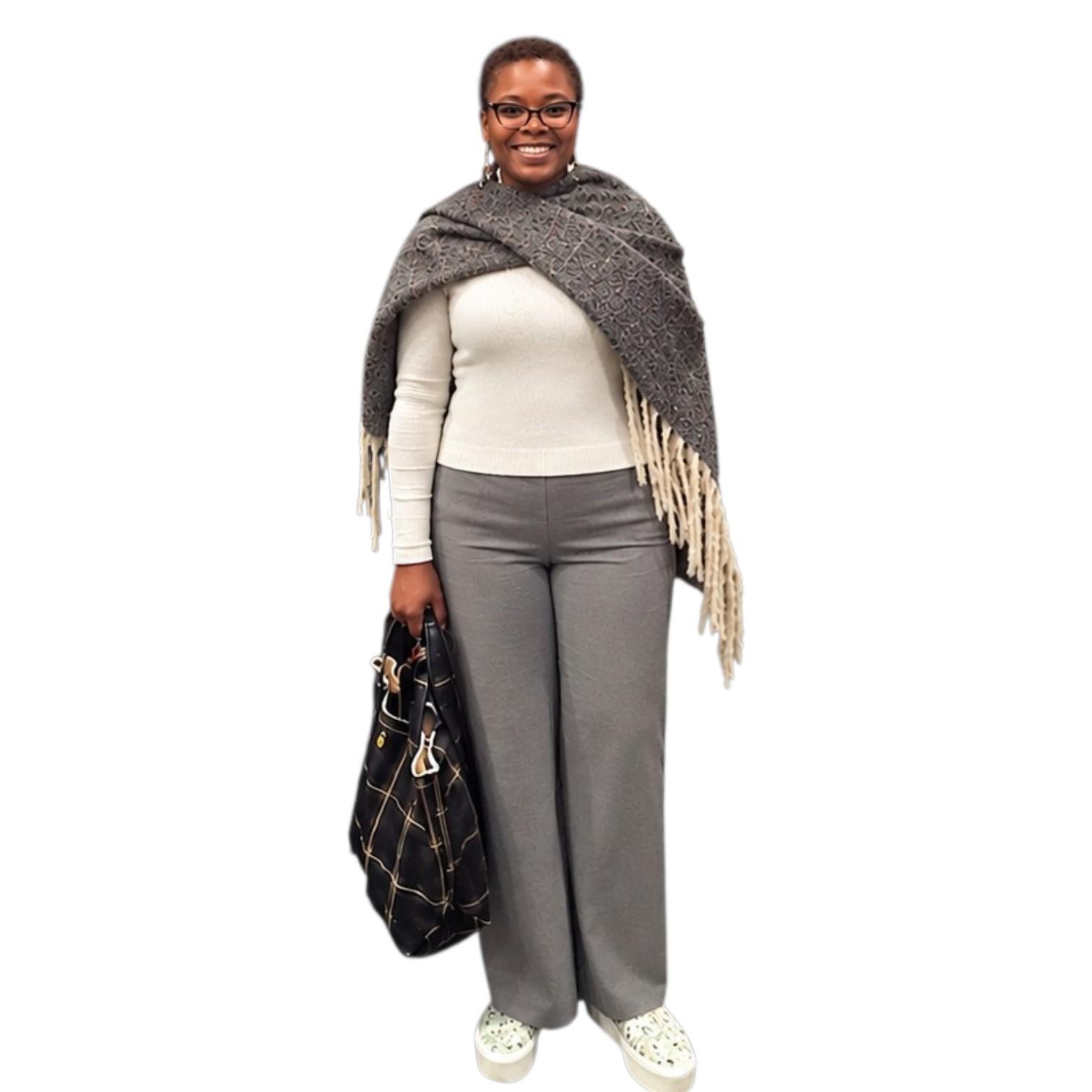 Woman smiling, wearing a gray shawl, white top, gray pants, and carrying a black bag