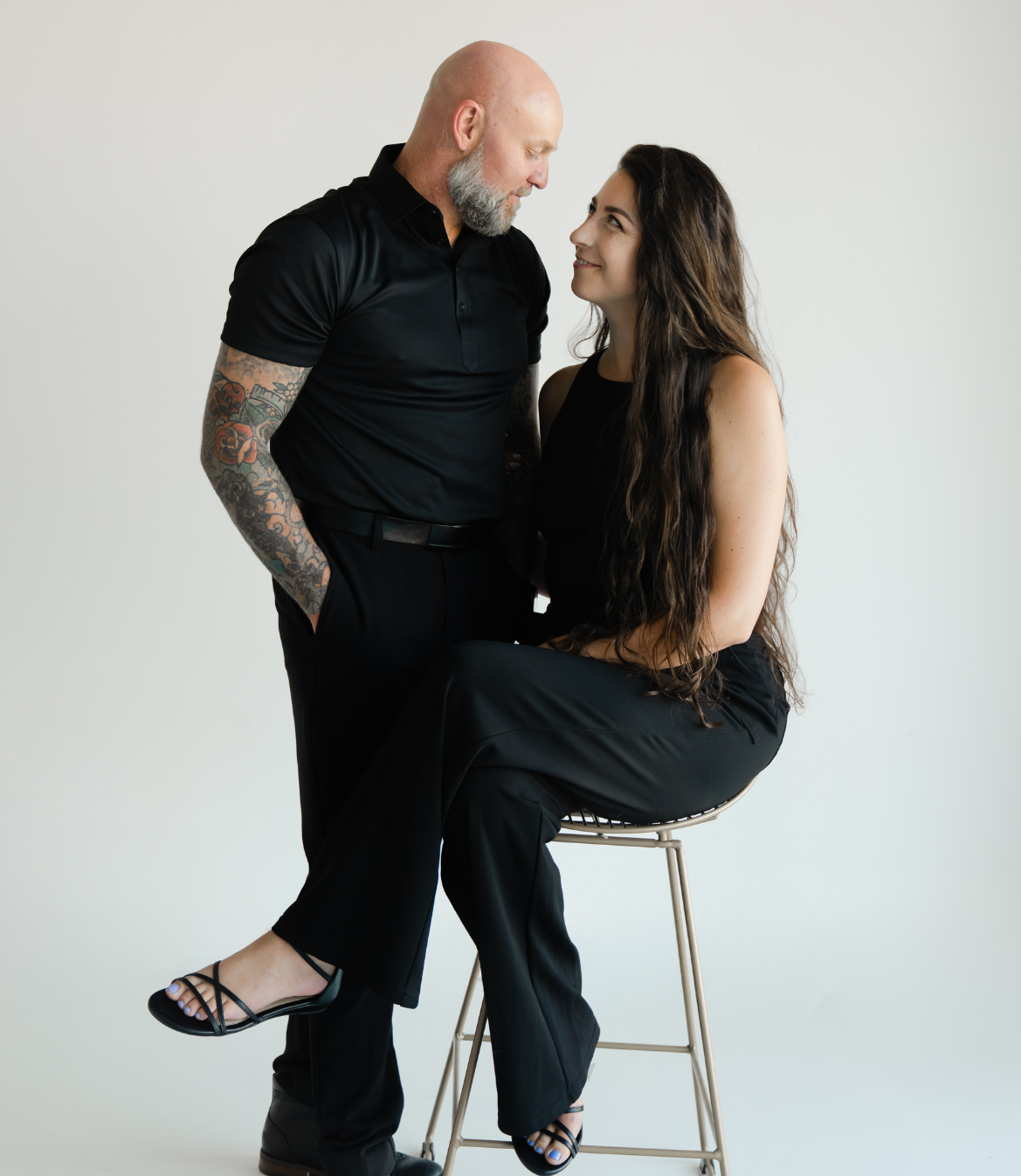 A couple in black clothing gazes at each other, sitting on a stool