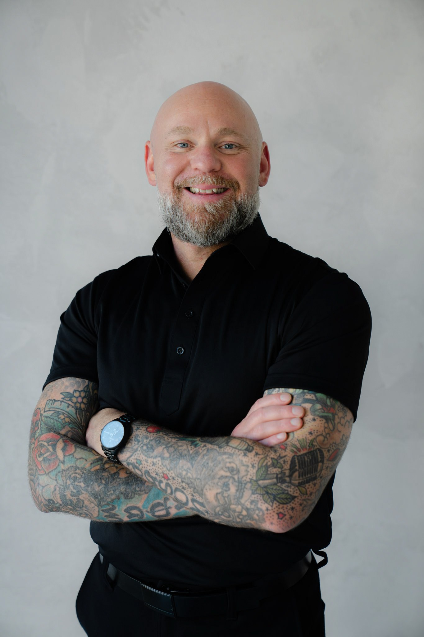 Smiling man with tattoos wearing a black polo shirt, arms crossed confidently