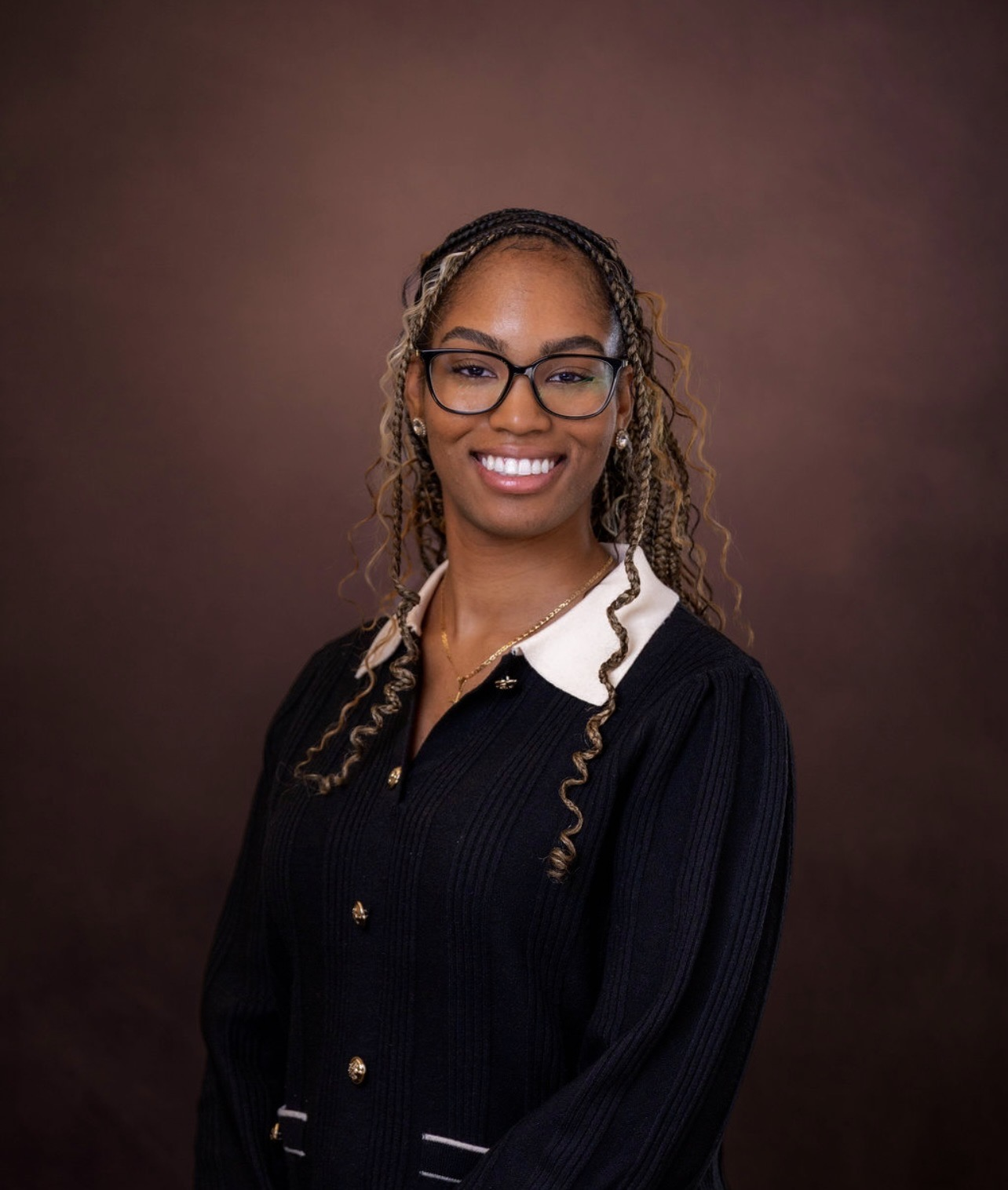 A smiling woman with curly hair wears glasses and a black sweater, against a brown background