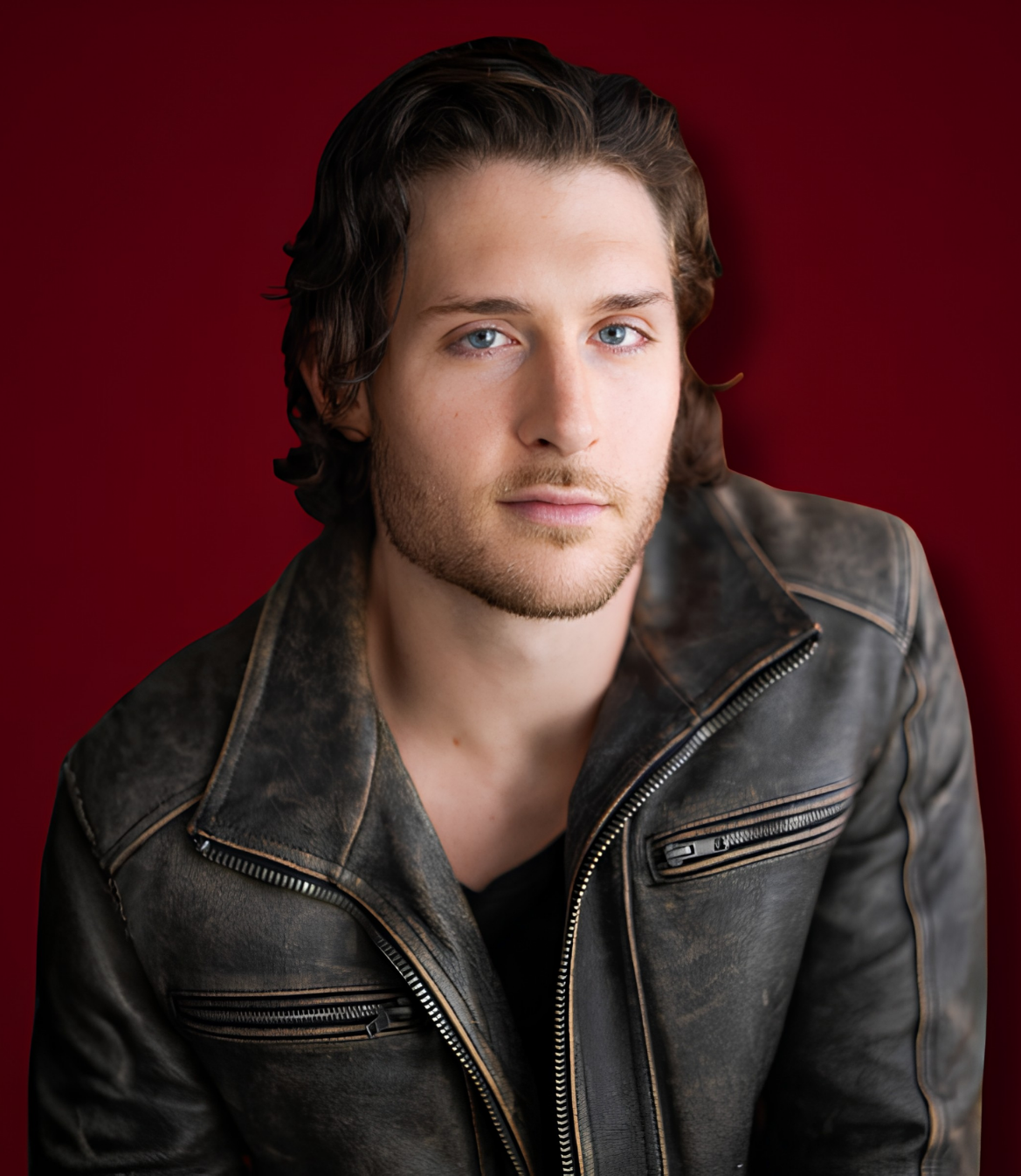Man with wavy hair wearing a leather jacket against a deep red background