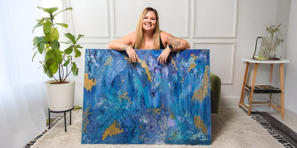 A woman smiles while holding a large, colorful abstract painting in a bright room