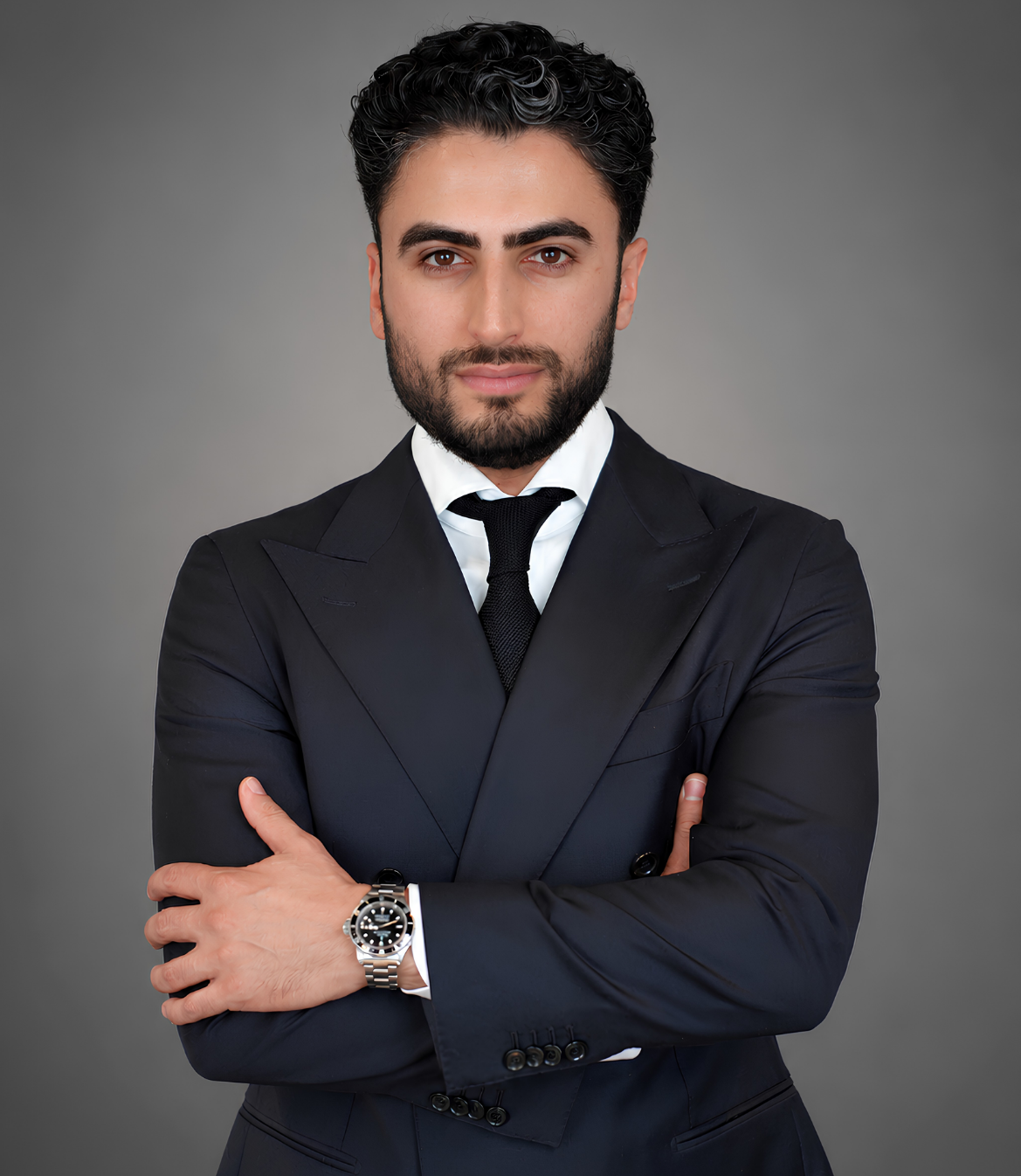 A well-dressed man with curly hair standing confidently with arms crossed.