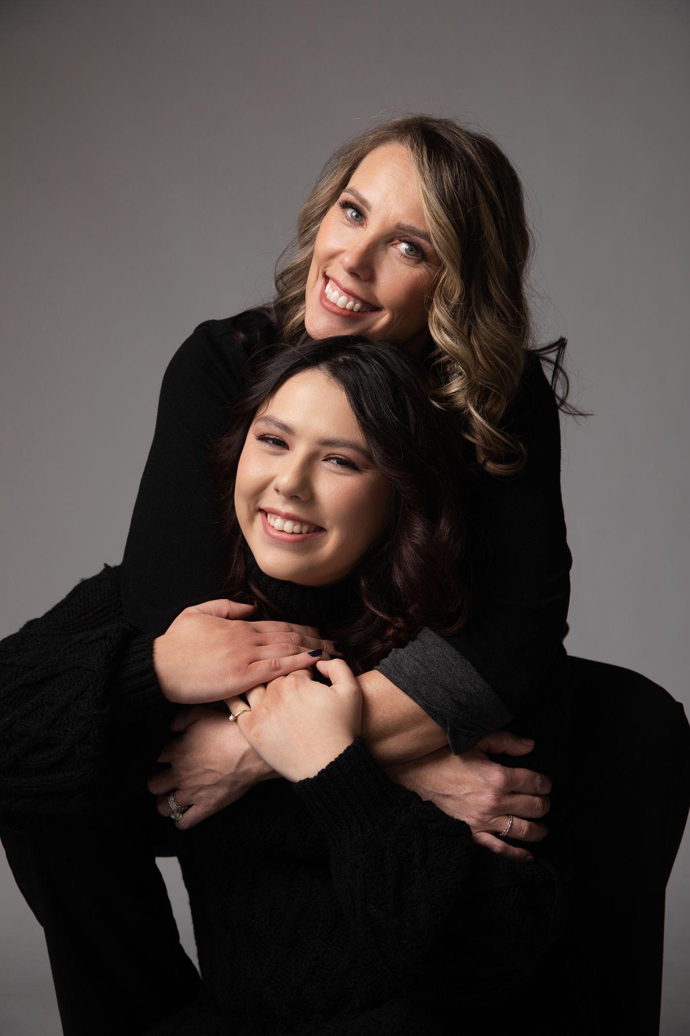 Two women embrace each other, smiling, in a studio setting with a gray background