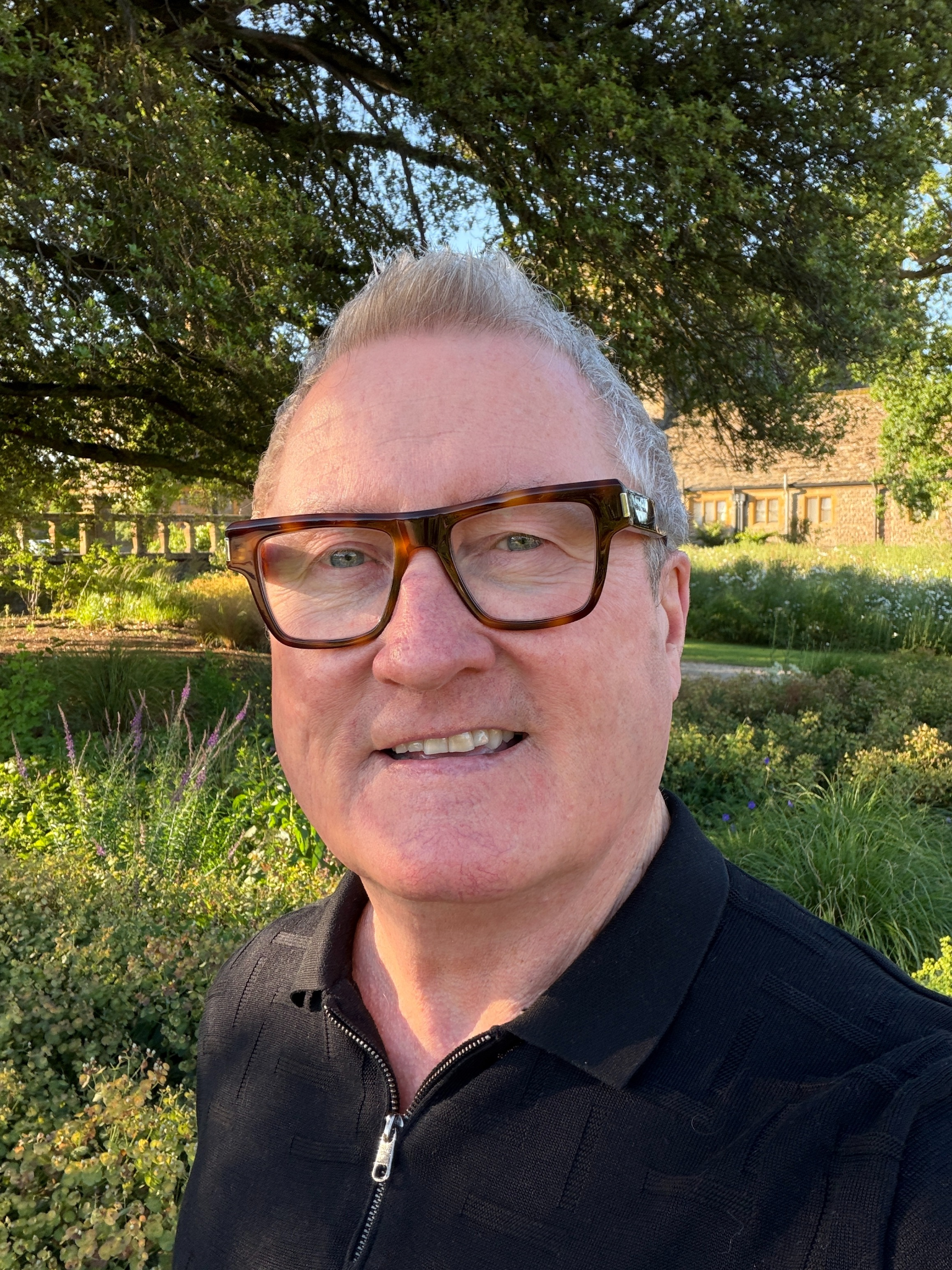Man with glasses smiling in a garden setting with greenery and flowers