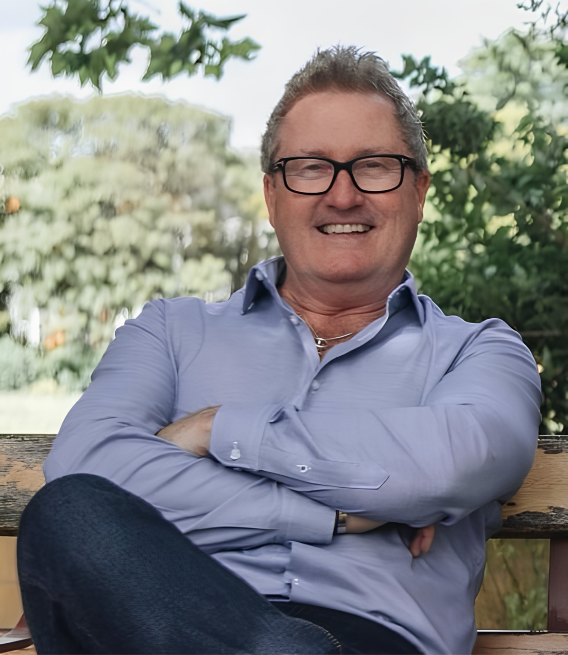 Smiling man in a blue shirt sitting on a bench surrounded by greenery