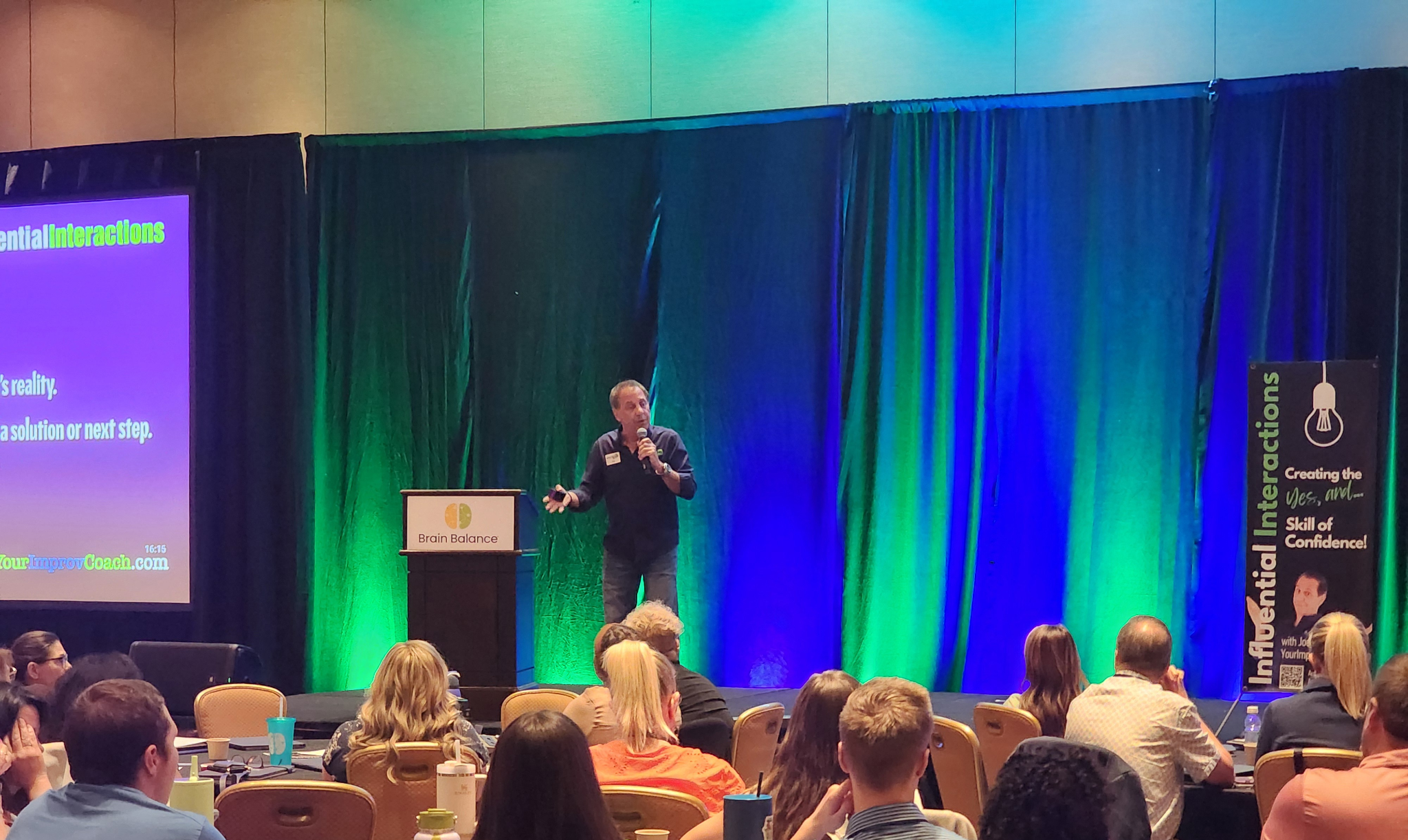A speaker addresses an audience at a conference, with colorful curtains in the background