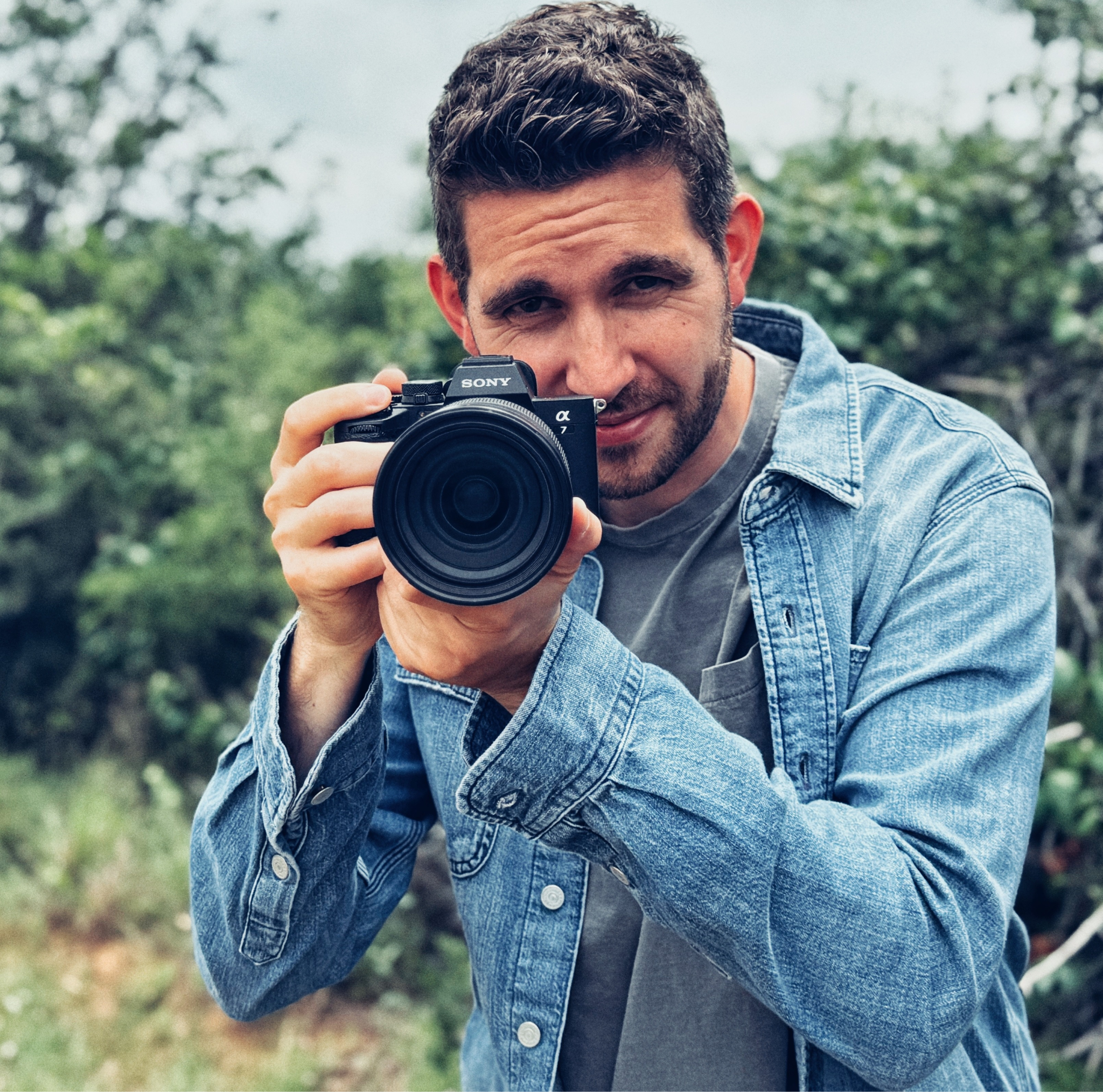A man holding a camera, wearing a denim jacket, outdoors with greenery behind.