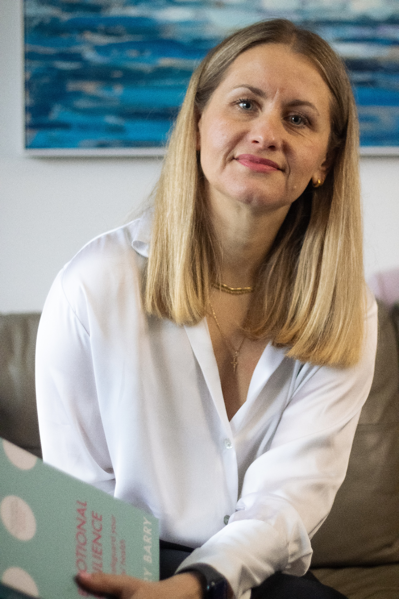 A woman with blonde hair sits on a couch holding a book, smiling at the camera