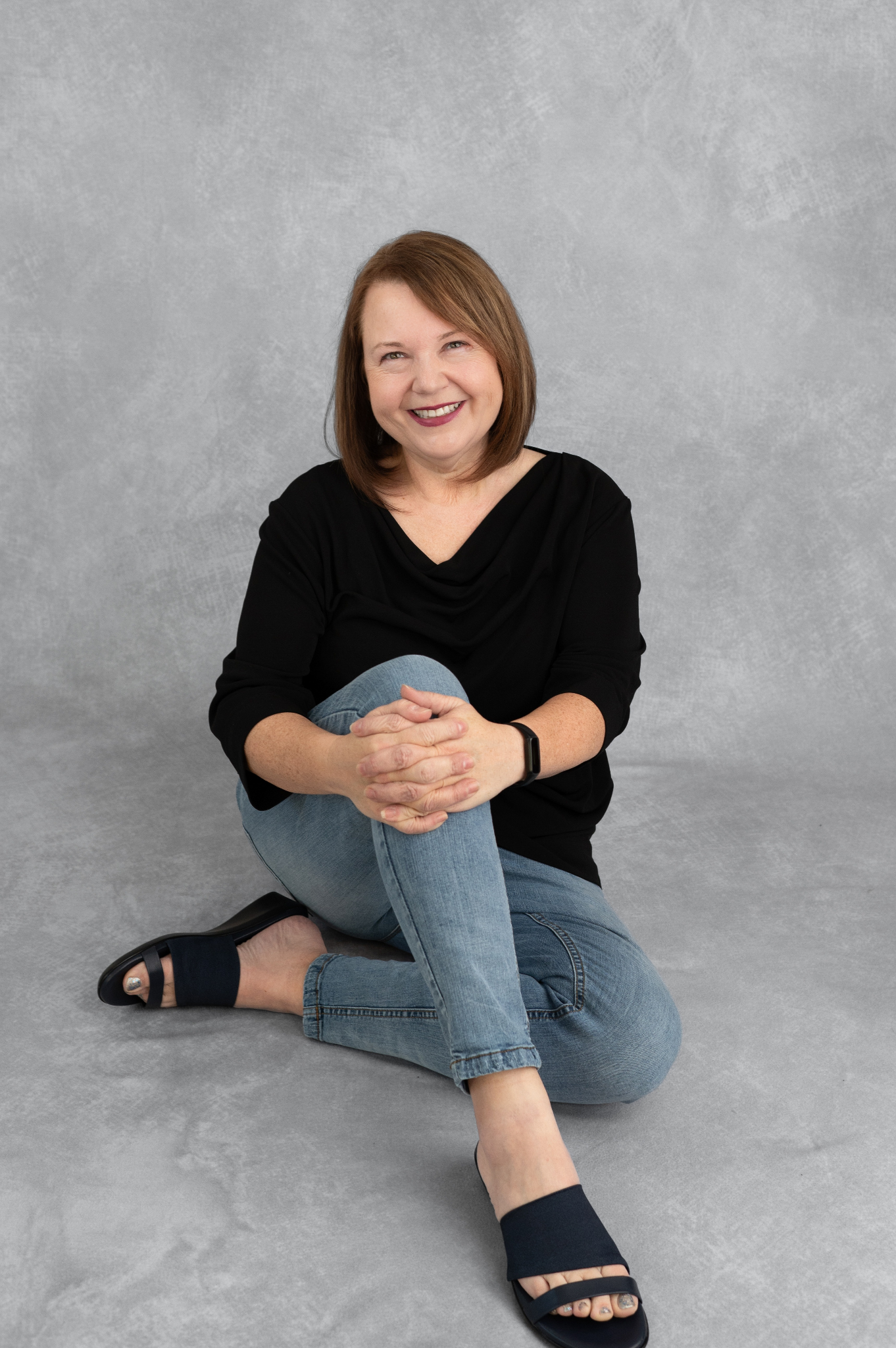 Smiling woman seated on the floor, wearing a black top and jeans