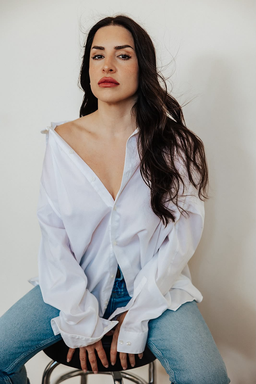 Woman in a white shirt seated on a stool, posing confidently against a plain background
