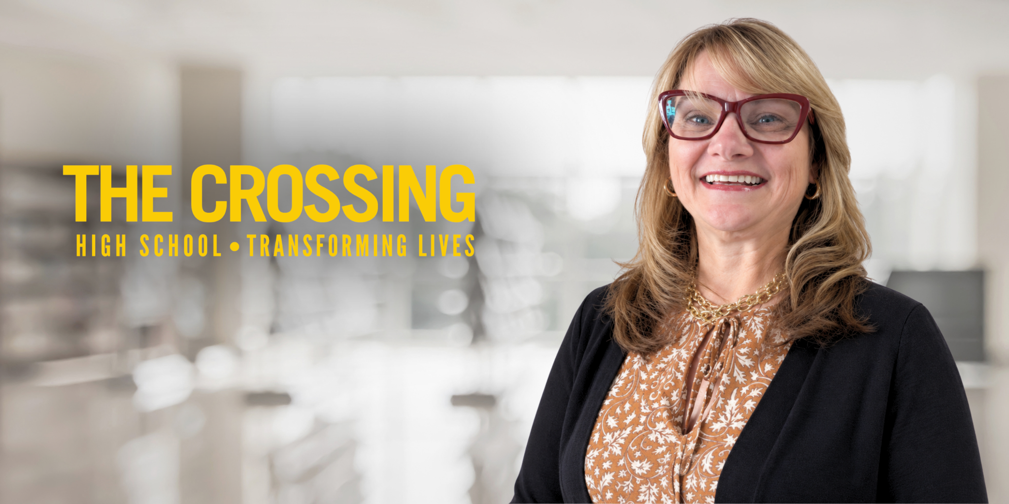 Smiling woman in glasses stands in a school setting with "The Crossing" logo