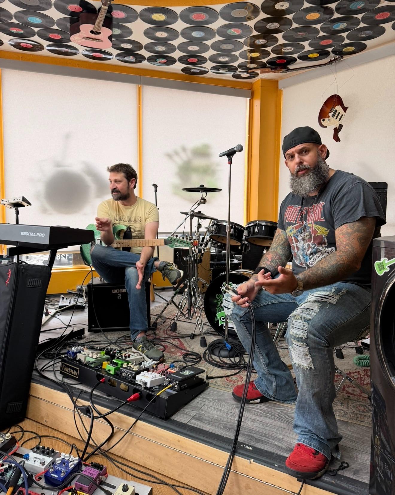 Two musicians seated in a studio, one playing guitar and the other on keyboard