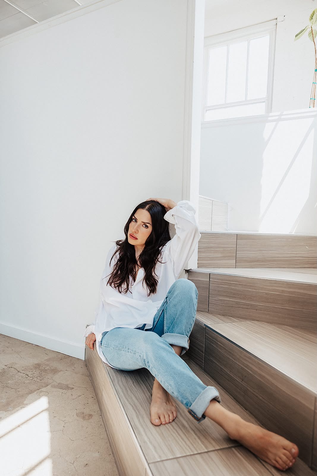 A woman in a white shirt and jeans sits on stairs, looking thoughtfully at the camera