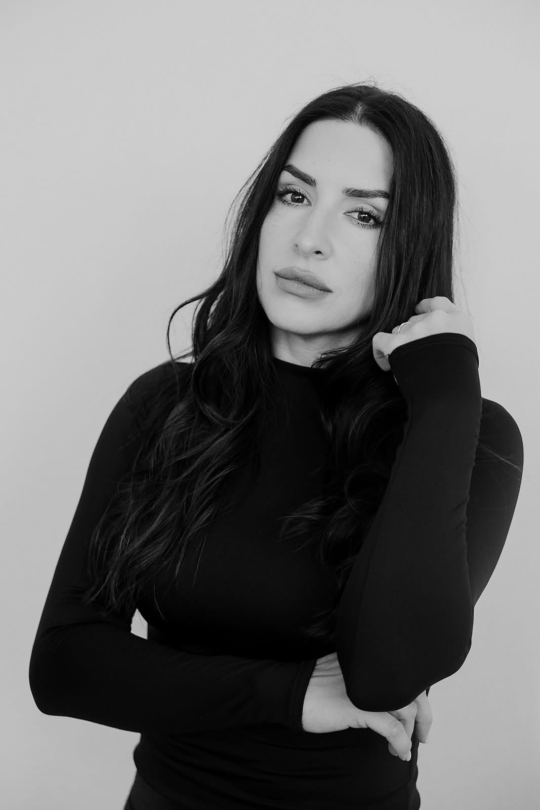A woman with long black hair poses thoughtfully in a black long-sleeve top