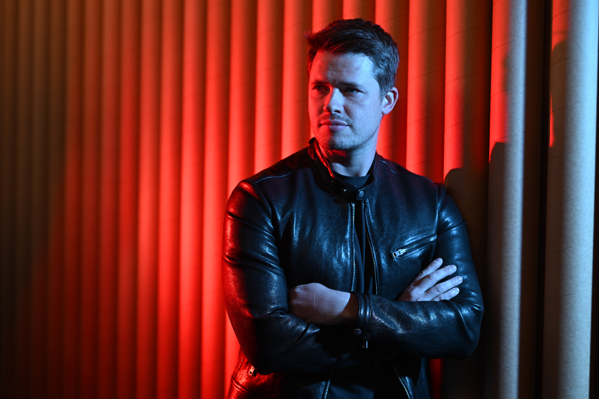 Man in a black leather jacket stands against a striped backdrop with red lighting