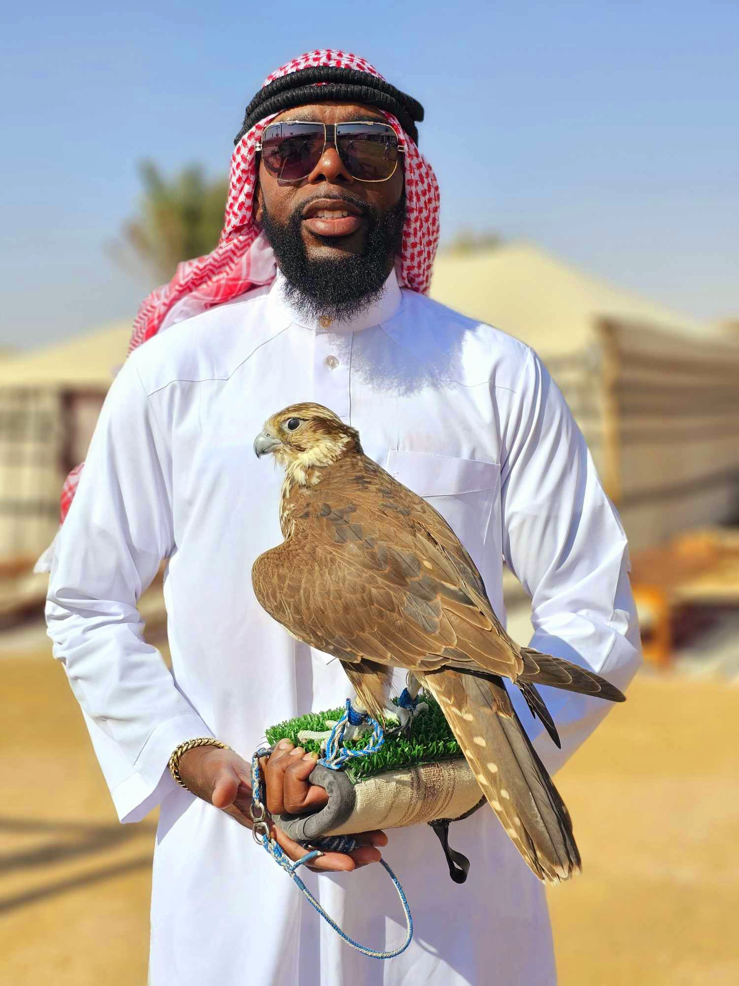 Man in traditional attire holding a falcon on a perch outdoors.