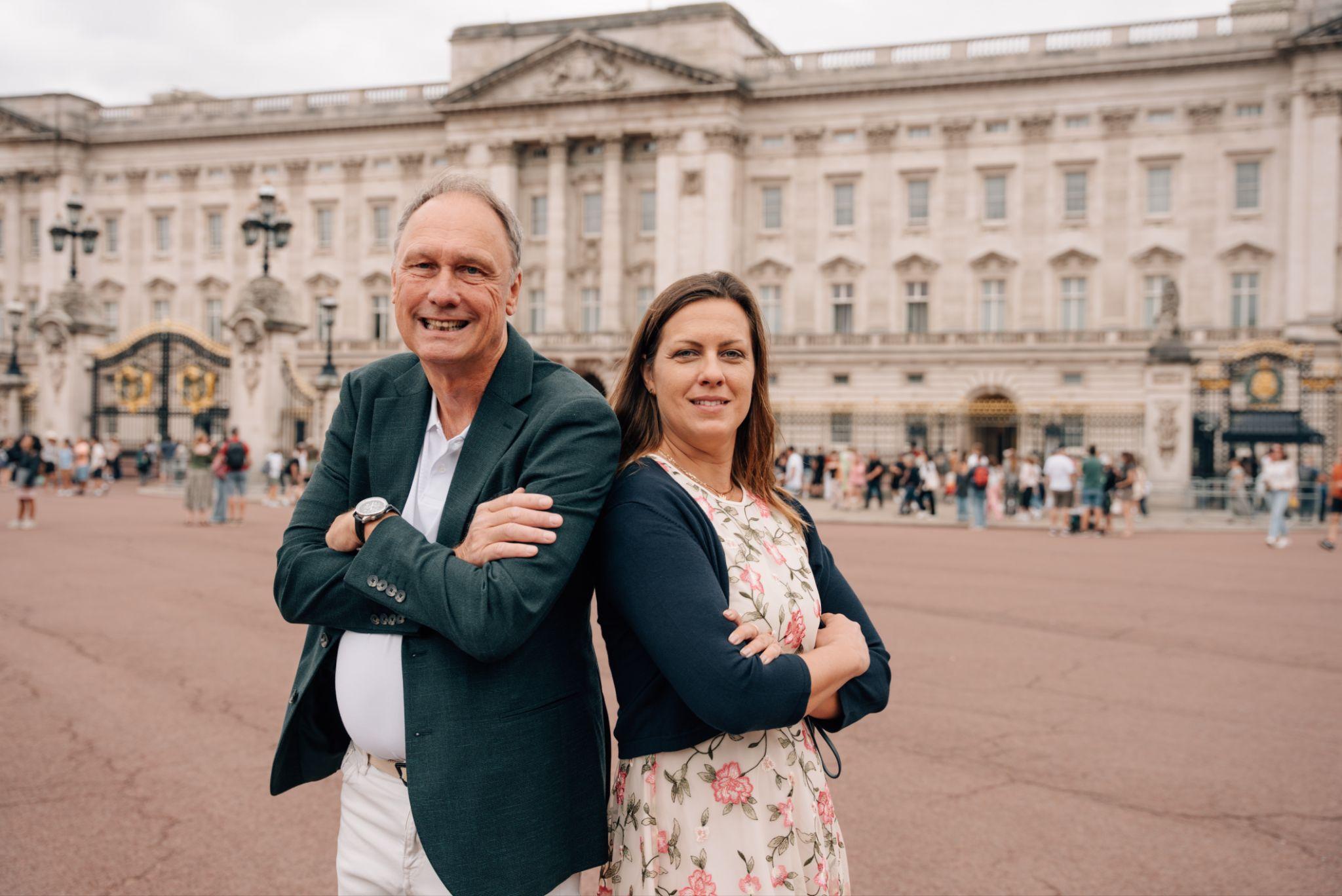 Two people pose in front of Buckingham Palace with arms crossed, smiling.