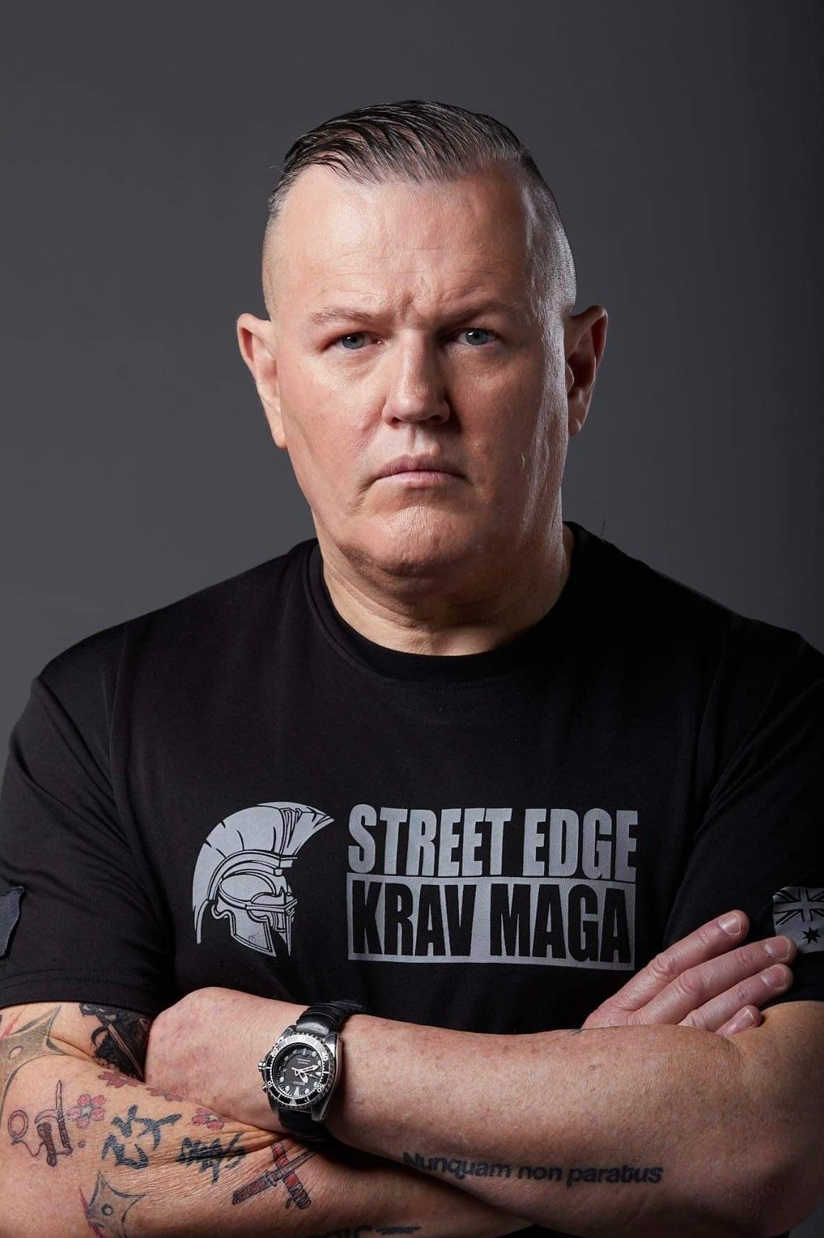A serious man with tattoos wearing a Krav Maga shirt, arms crossed.
