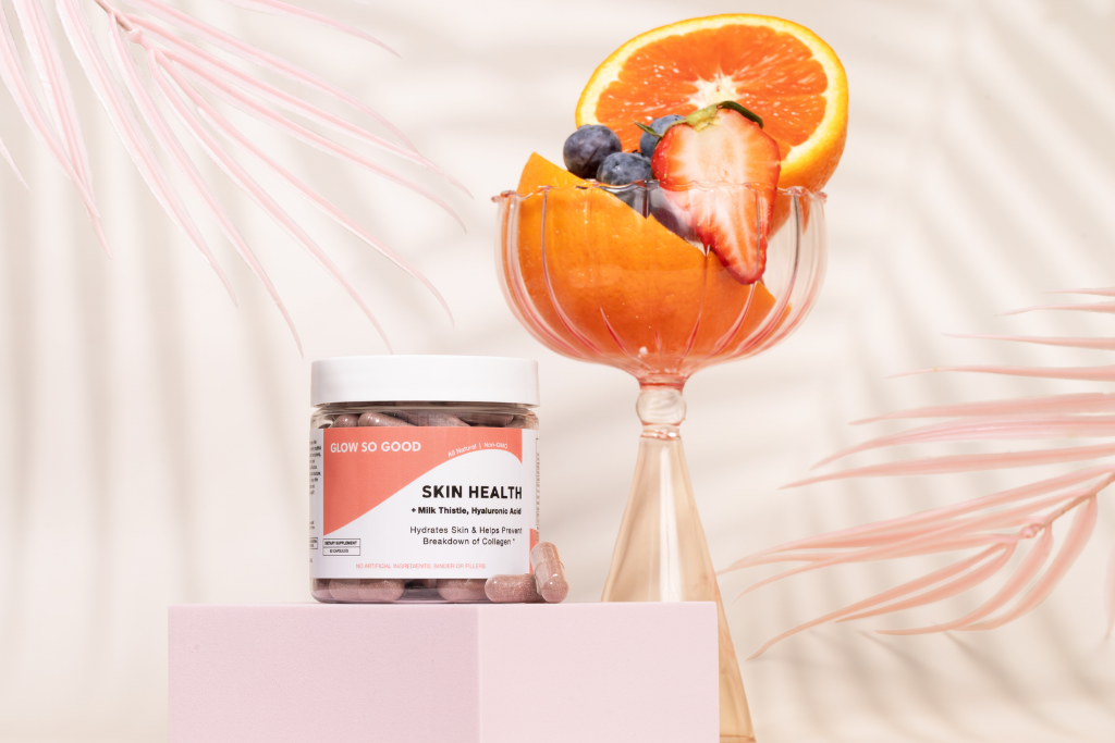 Vibrant fruit in a glass bowl beside a skincare supplement jar on a pink pedestal