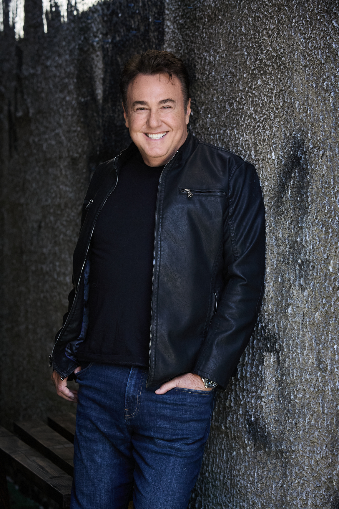 Man in a black leather jacket smiling against a textured gray background