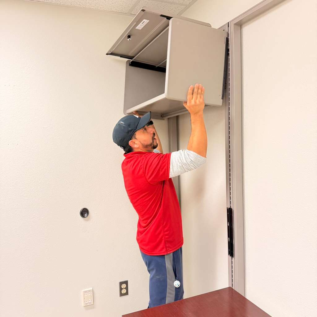 Man in red shirt lifting a gray cabinet against a wall in an office.