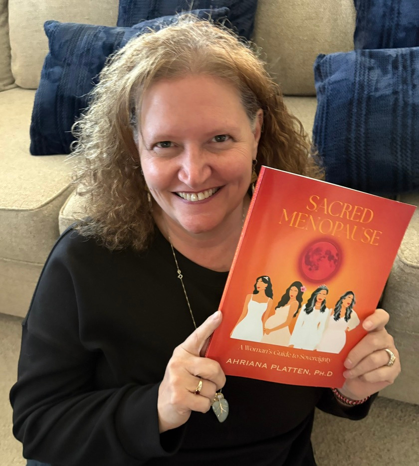 Smiling woman holding the book "Sacred Menopause" by Ahriana Platten on a couch