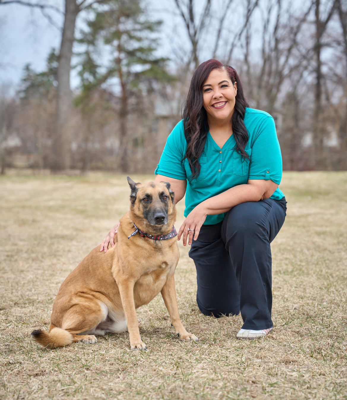 A woman kneels beside a tan dog in a grassy outdoor setting.