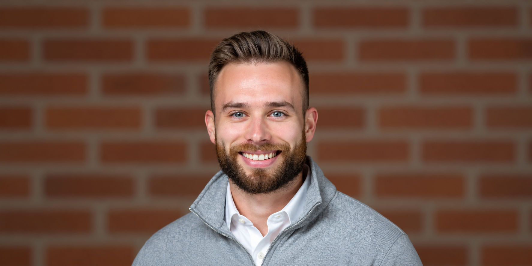 Smiling man with a beard wearing a gray sweater and white shirt against brick wall