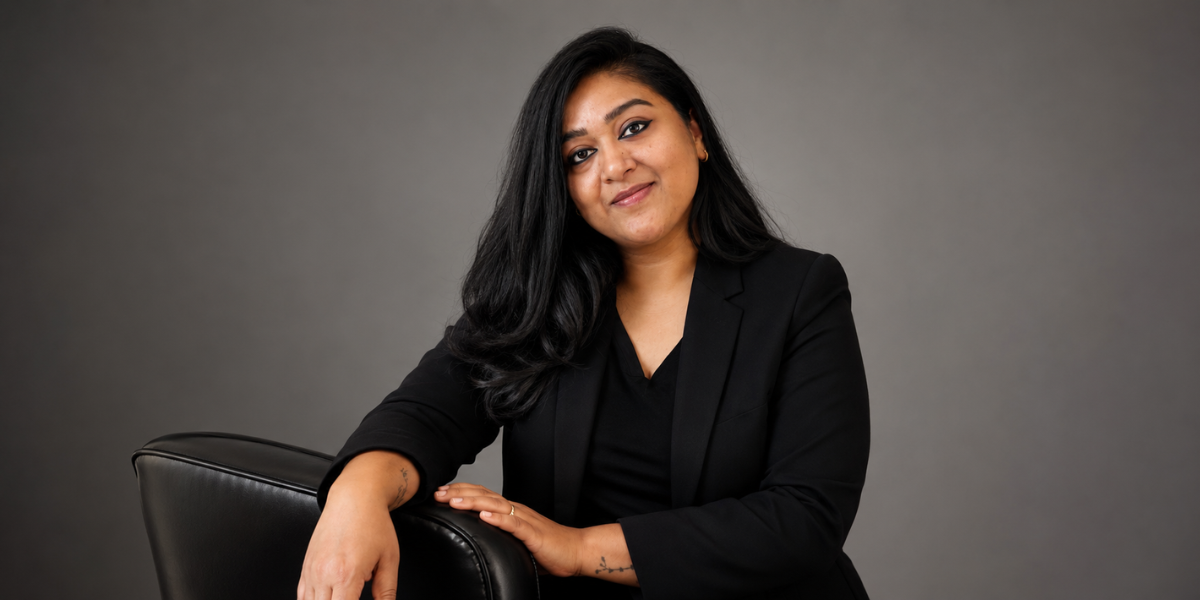 A woman in a black blazer poses confidently beside a chair against a gray backdrop